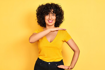 Young arab woman with curly hair wearing t-shirt standing over isolated yellow background gesturing with hands showing big and large size sign, measure symbol. Smiling looking at the camera. 