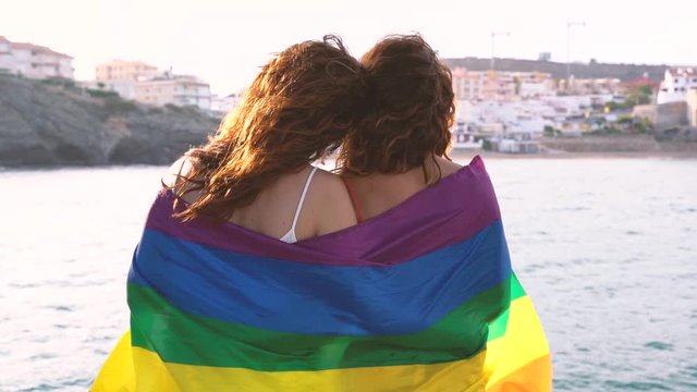 Tender lesbian couple staring at the sunset in the beach. Wrapped with a pride flag to show their love.Homosexual rights to have recognition in modern lifestyle. New generation free of prejudices.