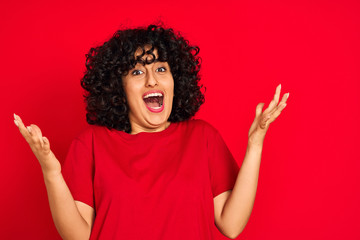 Young arab woman with curly hair wearing casual t-shirt over isolated red background celebrating crazy and amazed for success with arms raised and open eyes screaming excited. Winner concept