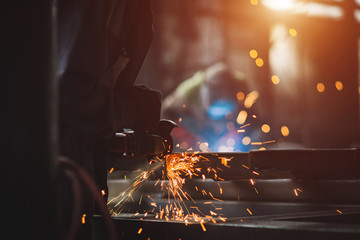 A man grinds metal. In the background a welder welds a part.