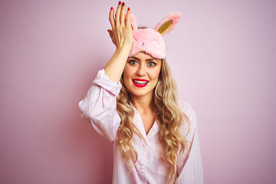 Young beautiful woman wearing pajama and sleep mask over pink isolated background surprised with hand on head for mistake, remember error. Forgot, bad memory concept.
