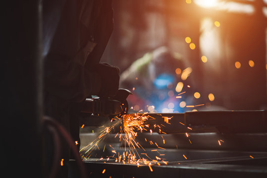 A man grinds metal. In the background a welder welds a part.