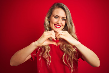 Young beautiful woman wearing basic t-shirt standing over red isolated background smiling in love doing heart symbol shape with hands. Romantic concept.