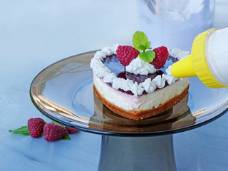 Decorating a heart shaped cheesecake  with whipped cream. No-bake cheesecake with graham cracker crust, cream cheese filling and raspberry jelly on glass cake stand.