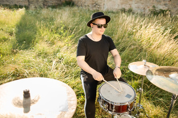 Drummer in black hat rehearses in the open air. He plays drums in the grass near the old castle