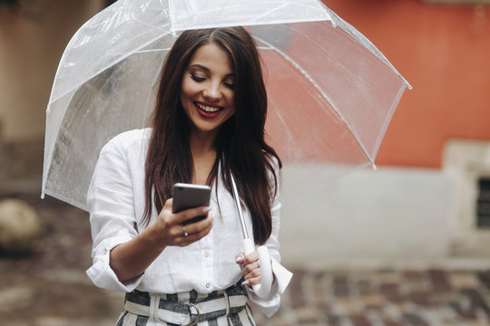 Portrait of smiling beautiful girl with umbrella. using smartphone, red house on the background. It's summer rain.