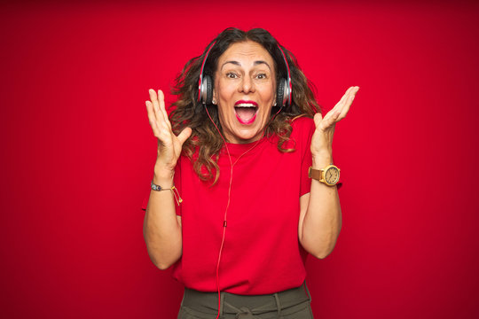 Middle age senior woman wearing headphones listening to music over red isolated background celebrating crazy and amazed for success with arms raised and open eyes screaming excited. Winner concept