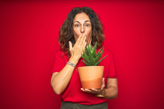 Middle age senior woman holding green cactus over red isolated background cover mouth with hand shocked with shame for mistake, expression of fear, scared in silence, secret concept
