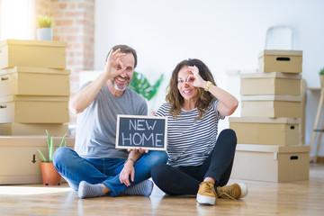 Fototapeta na wymiar Middle age senior couple sitting on the floor holding blackboard moving to a new home with happy face smiling doing ok sign with hand on eye looking through fingers