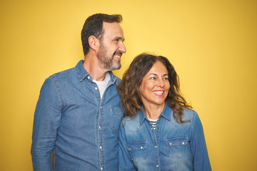 Beautiful middle age couple together standing over isolated yellow background looking away to side with smile on face, natural expression. Laughing confident.