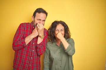 Beautiful middle age couple over isolated yellow background smelling something stinky and...