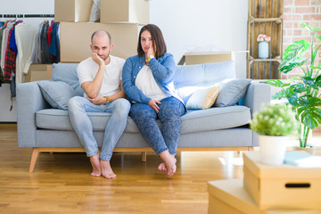Young couple sitting on the sofa arround cardboard boxes moving to a new house thinking looking tired and bored with depression problems with crossed arms.