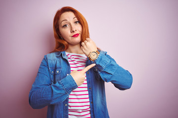 Beautiful redhead woman wearing denim shirt and striped t-shirt over isolated pink background In hurry pointing to watch time, impatience, looking at the camera with relaxed expression