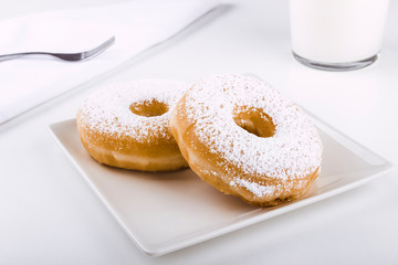 Fototapeta na wymiar Donuts covered with icing sugar on a plate on a white background accompanied by a glass of milk and a fork on a napkin. Delicious breakfast or lunch. American food. Bakery and pastry products.