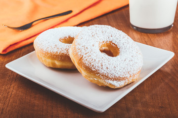 Donuts covered with icing sugar on a plate on a wooden table accompanied by a glass of milk and a fork on an orange napkin. Delicious breakfast or lunch. American food. Bakery and pastry products.