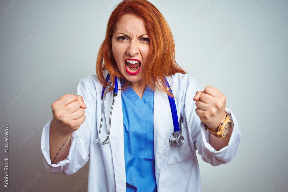 Sticker Young redhead doctor woman using stethoscope over white isolated background angry and mad raising fists frustrated and furious while shouting with anger. Rage and aggressive concept.