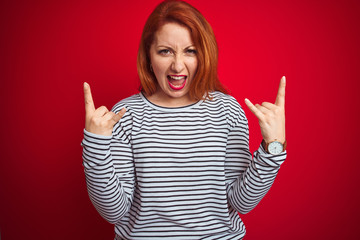 Young redhead woman wearing strapes navy shirt standing over red isolated background shouting with crazy expression doing rock symbol with hands up. Music star. Heavy concept.