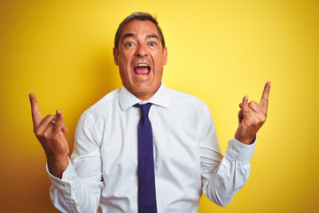 Handsome middle age businessman standing over isolated yellow background shouting with crazy expression doing rock symbol with hands up. Music star. Heavy concept.