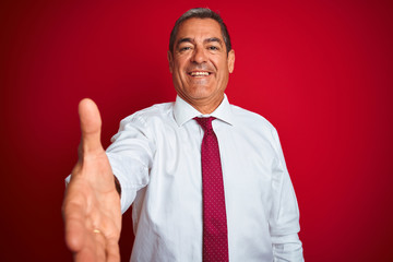 Handsome middle age businessman standing over isolated red background smiling friendly offering handshake as greeting and welcoming. Successful business.