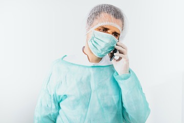 The surgeon speaks on the phone, consults wearing a green surgical apron and a face mask on a light background. Medical and pharmaceutical concept. The surgeon needs the help of another doctor.
