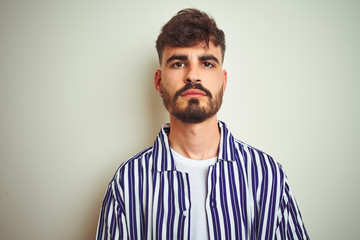 Young man with tattoo wearing striped shirt standing over isolated white background with serious expression on face. Simple and natural looking at the camera.