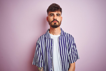 Young man with tattoo wearing striped shirt standing over isolated pink background Relaxed with serious expression on face. Simple and natural looking at the camera.