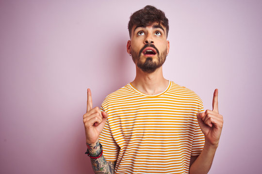 Young man with tattoo wearing yellow striped t-shirt standing over isolated pink background amazed and surprised looking up and pointing with fingers and raised arms.