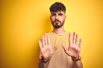 Young man with tattoo wearing striped t-shirt standing over isolated yellow background Moving away...