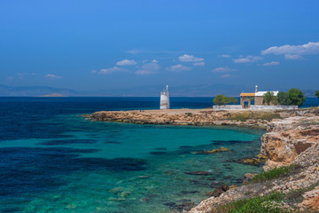Fototapeta na wymiar The wild coast of Aegina island with clear and blue waters of Mediterranean sea and the old small lighthouse in the background, in Saronic gulf, Greece.