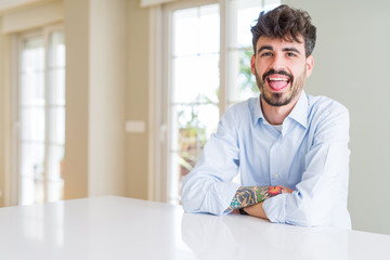 Fototapeta premium Young businesss man sitting on white table sticking tongue out happy with funny expression. Emotion concept.