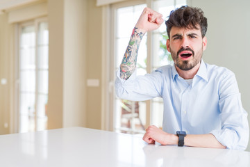 Young businesss man sitting on white table angry and mad raising fist frustrated and furious while shouting with anger. Rage and aggressive concept.