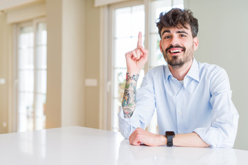 Fototapeta premium Young businesss man sitting on white table pointing finger up with successful idea. Exited and happy. Number one.