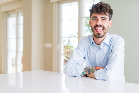 Young businesss man sitting on white table sticking tongue out happy with funny expression. Emotion concept.