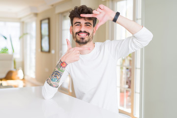 Fototapeta premium Young man wearing casual shirt sitting on white table smiling making frame with hands and fingers with happy face. Creativity and photography concept.