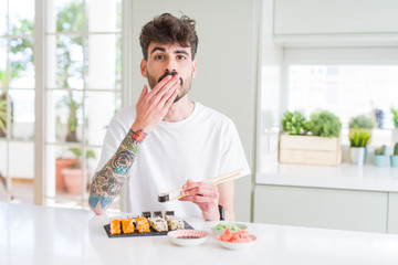 Young man eating asian sushi from home delivery cover mouth with hand shocked with shame for mistake, expression of fear, scared in silence, secret concept