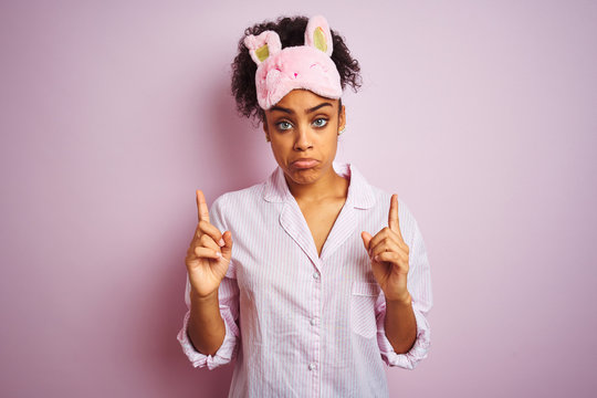 Young african american woman wearing pajama and mask over isolated pink background Pointing up looking sad and upset, indicating direction with fingers, unhappy and depressed. - Powered by Adobe