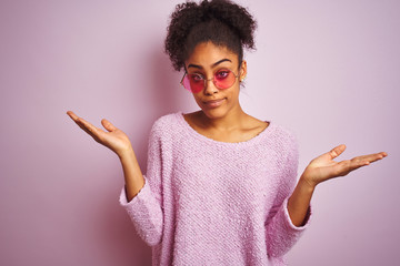 African american woman wearing winter sweater and sunglasses over isolated pink background clueless and confused expression with arms and hands raised. Doubt concept.