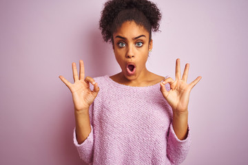 Young african american woman wearing winter sweater standing over isolated pink background looking surprised and shocked doing ok approval symbol with fingers. Crazy expression