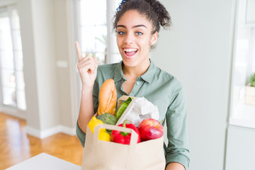 Young african american girl holding paper bag of groceries from supermarket very happy pointing with hand and finger to the side