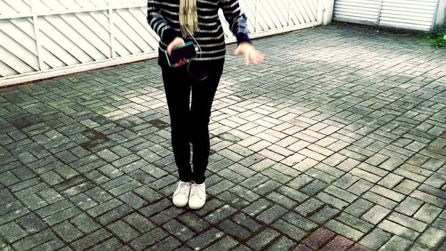 Funny girl dancing and listening music on cellphone on white gate background. Unrecognizable model body holding smartphone and earphones wearing casual black pants and pullover and white sneakers.