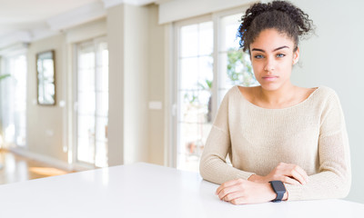 Beautiful young african american woman with afro hair sitting on table at home skeptic and nervous,...