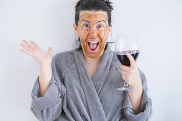 Beautiful woman wearing cosmetic facial mask as skincare treatment drinking glass of wine very happy and excited, winner expression celebrating victory screaming with big smile and raised hands