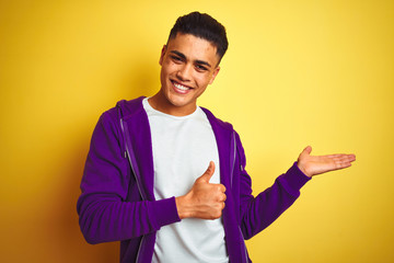 Young brazilian man wearing purple sweatshirt standing over isolated yellow background Showing palm hand and doing ok gesture with thumbs up, smiling happy and cheerful