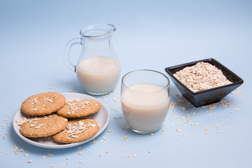 Leche de avena vegana sobre una mesa en un vaso de cristal, una jarra, un bol con copos de avena y un plato de galletas.
