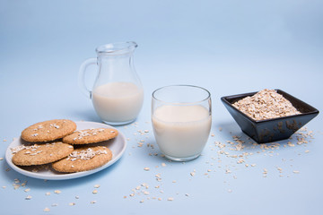 Leche de avena vegana sobre una mesa en un vaso de cristal, una jarra, un bol con copos de avena y un plato de galletas.	