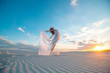 Fototapeta na wymiar A girl in a fly white dress dances and poses in the sand desert at sunset