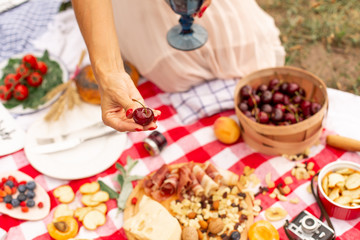 Girl holds juicy ripe cherries in her hand against the background of a checkered picnic blanket with food spread on it