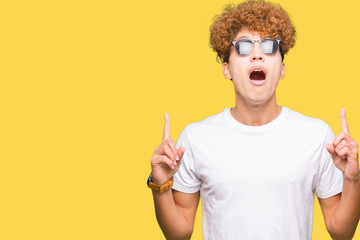 Young handsome man with afro hair wearing sunglasses amazed and surprised looking up and pointing with fingers and raised arms.
