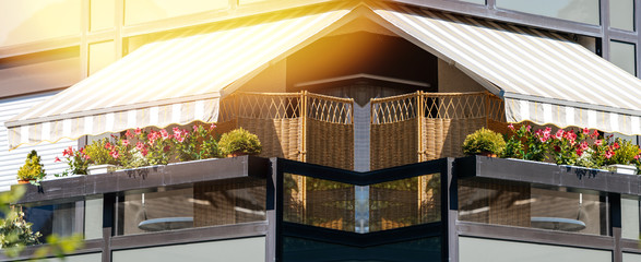 French long balcony with beautiful awning and flowers covered with rays of sun - protection during hot weather and radiation - sunlight flare - obrazy, fototapety, plakaty