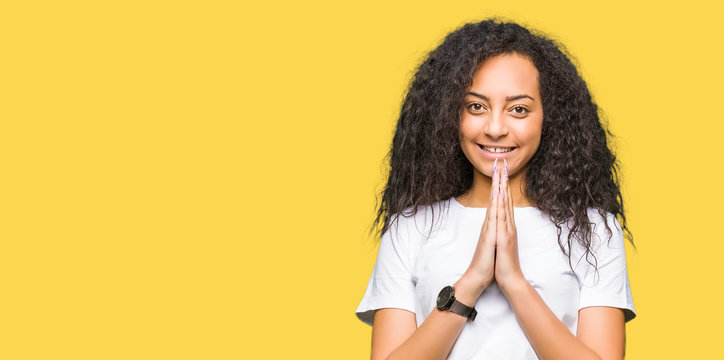 Young beautiful girl with curly hair wearing casual white t-shirt praying with hands together asking for forgiveness smiling confident.